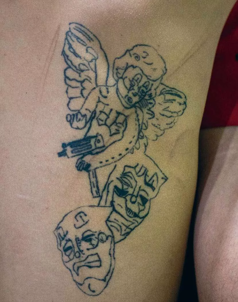 Un tatouage représentant un ange avec des ailes tenant une arme, au-dessus de trois masques de théâtre avec des expressions différentes.