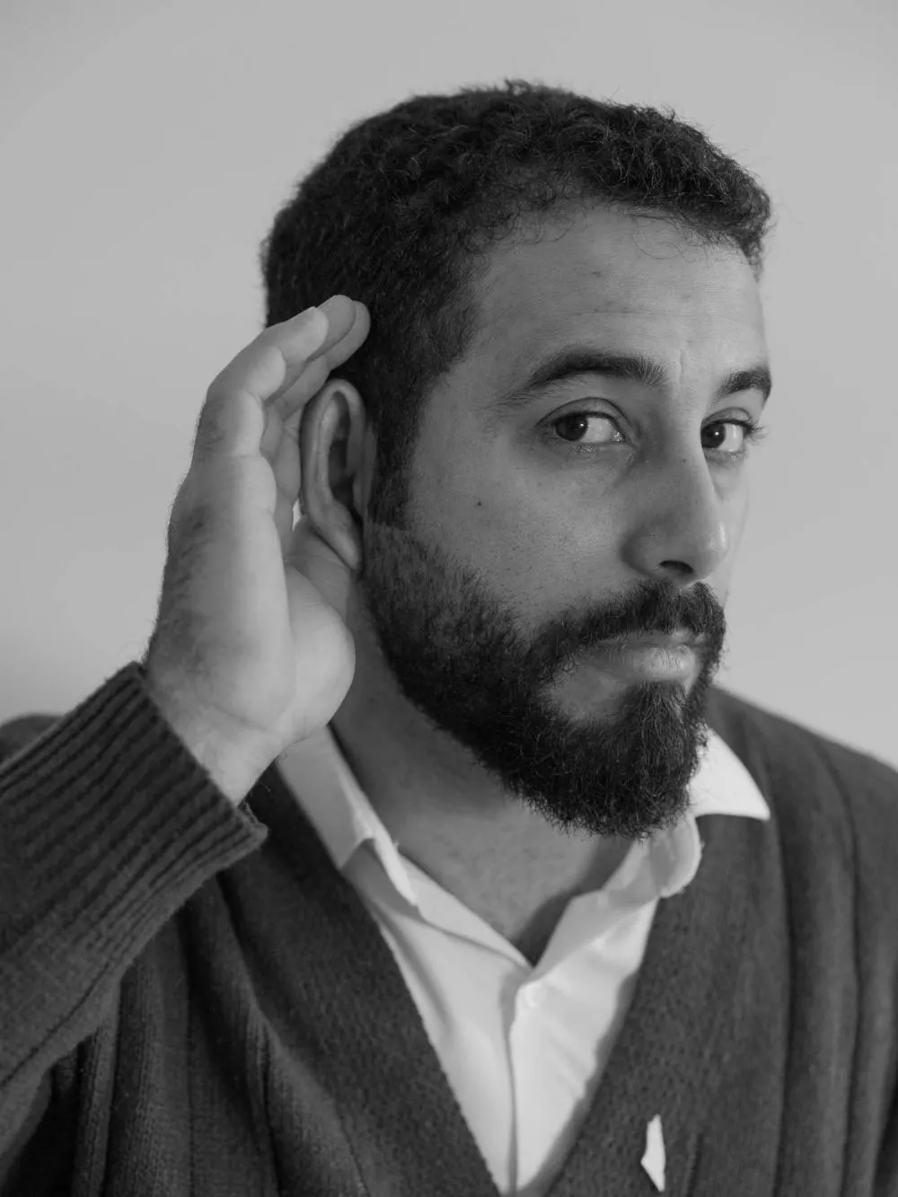 Un homme barbu en noir et blanc tient sa main près de son oreille, comme s'il écoutait attentivement. Il porte un pull et une chemise.