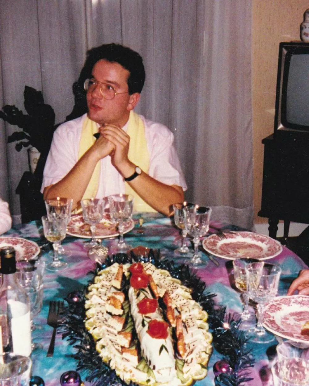 Un homme est assis à une table joliment dressée avec des plats et des verres. Il porte une chemise blanche et une écharpe jaune. Devant lui, il y a un plat décoré avec des aliments et des garnitures colorées.