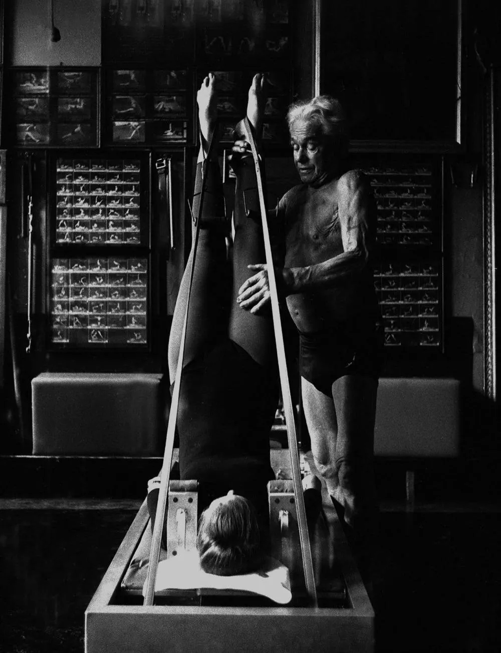Un homme âgé, torse nu, assiste une personne allongée sur un appareil de Pilates, les jambes en l'air, attachées par des sangles. L'environnement semble être un studio de fitness ou de rééducation.