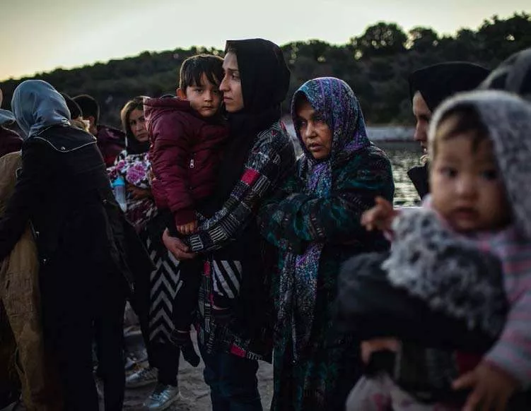 Un groupe de personnes, dont plusieurs femmes portant des foulards, se tient près de l'eau. Deux femmes tiennent des enfants dans leurs bras. L'atmosphère semble sérieuse et contemplative.