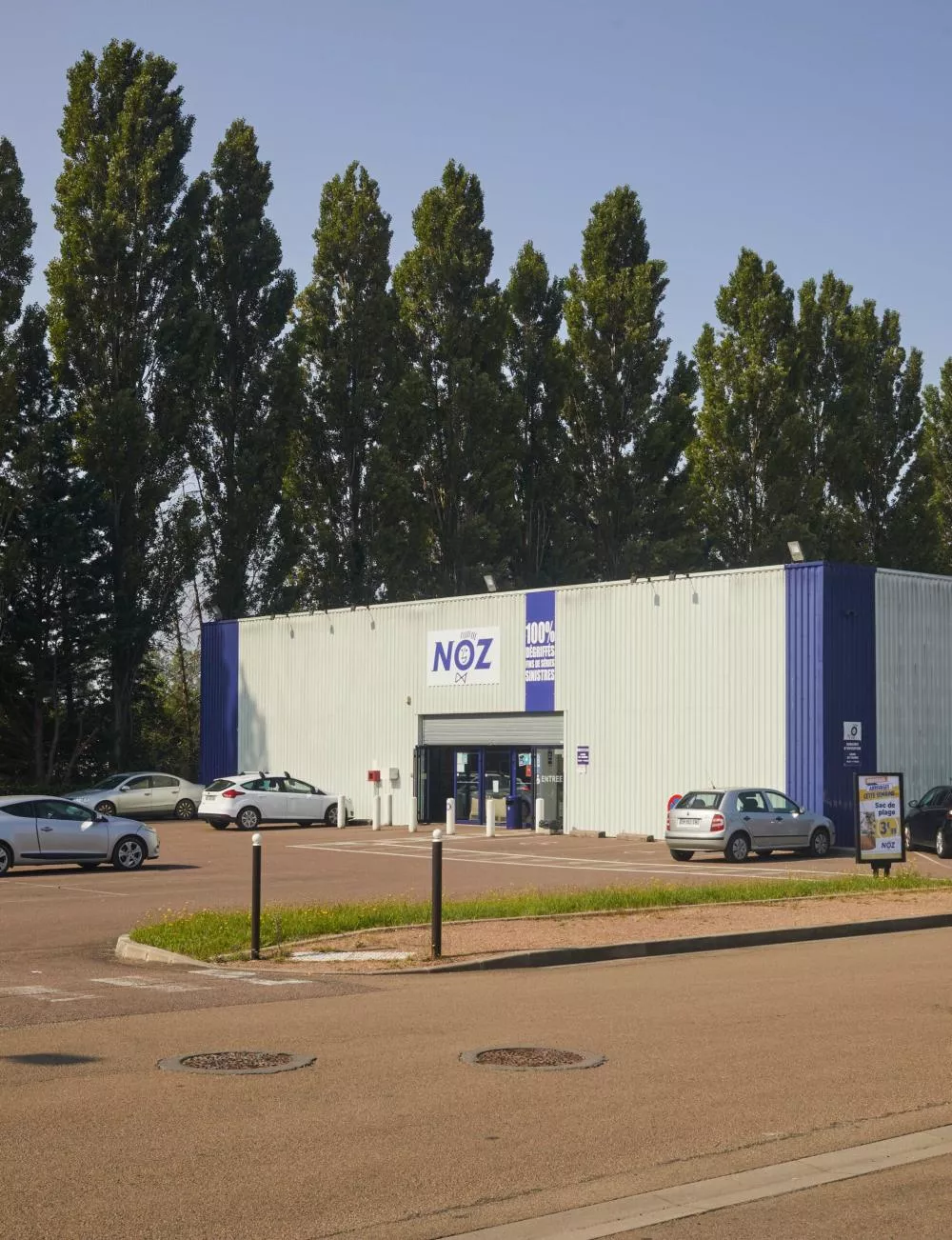 Un bâtiment avec le logo "NOZ" sur la façade, entouré de voitures garées et d'arbres en arrière-plan.