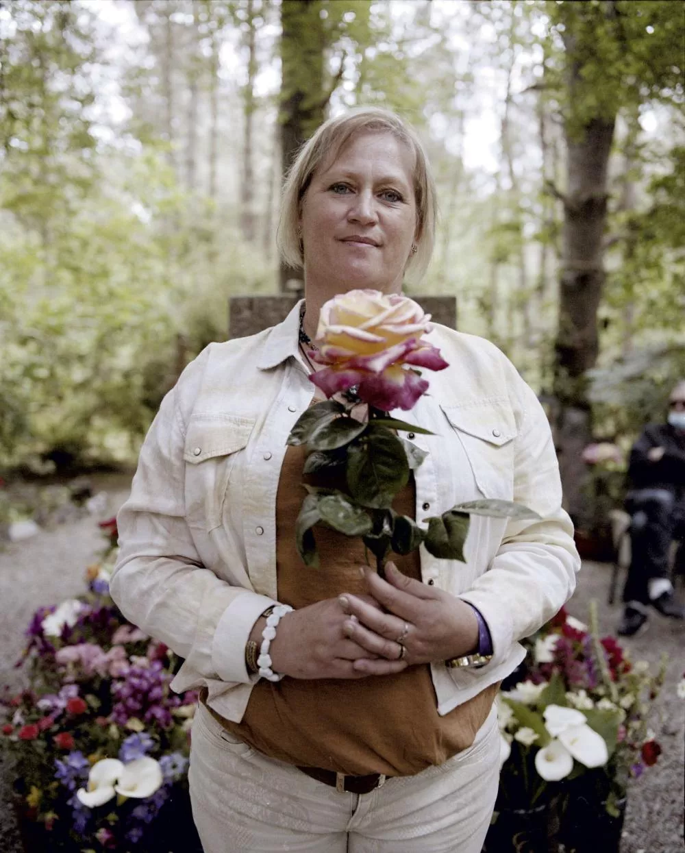 Une femme tient une rose multicolore dans un jardin ou un parc, entourée de fleurs et de verdure. Elle porte une veste claire et un bracelet blanc.