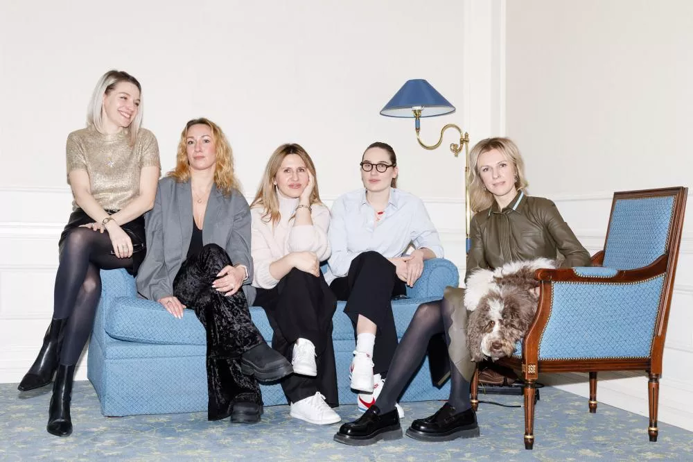 Cinq femmes sont assises dans un salon, quatre sur un canapé bleu et une sur un fauteuil avec un chien. Elles sourient et semblent détendues.
