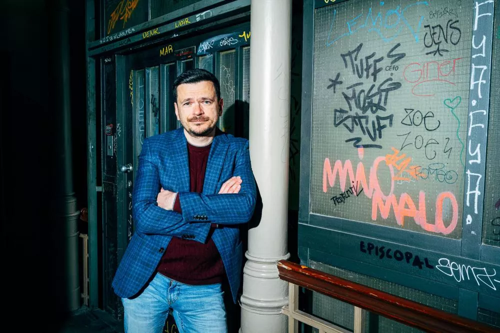 Un homme en veste bleue et pull bordeaux est debout, bras croisés, devant une porte métallique couverte de graffitis colorés.