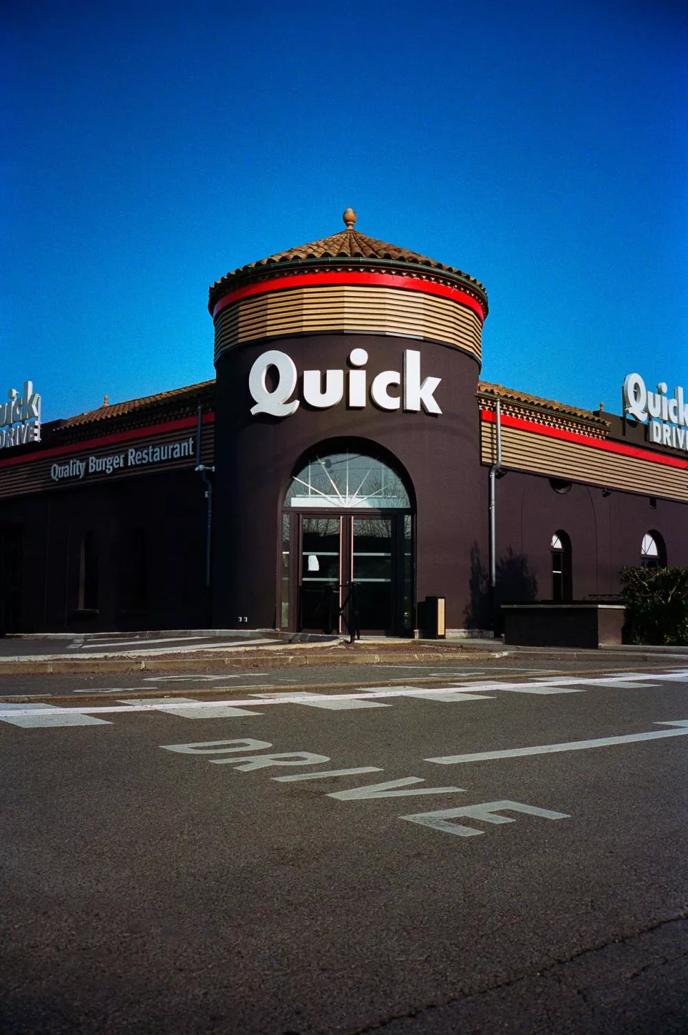 Un restaurant Quick avec une entrée principale visible, des enseignes indiquant "Quick Drive" et "Quality Burger Restaurant", sur un fond de ciel bleu.