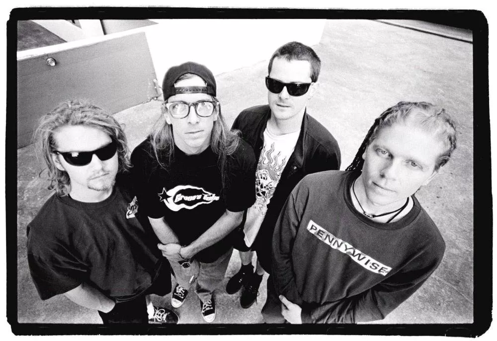 Quatre personnes portant des lunettes de soleil et des vêtements décontractés, regardant vers le haut. L'une porte une casquette à l'envers, une autre a des dreadlocks, et deux d'entre elles portent des t-shirts avec des logos. L'image est en noir et blanc.