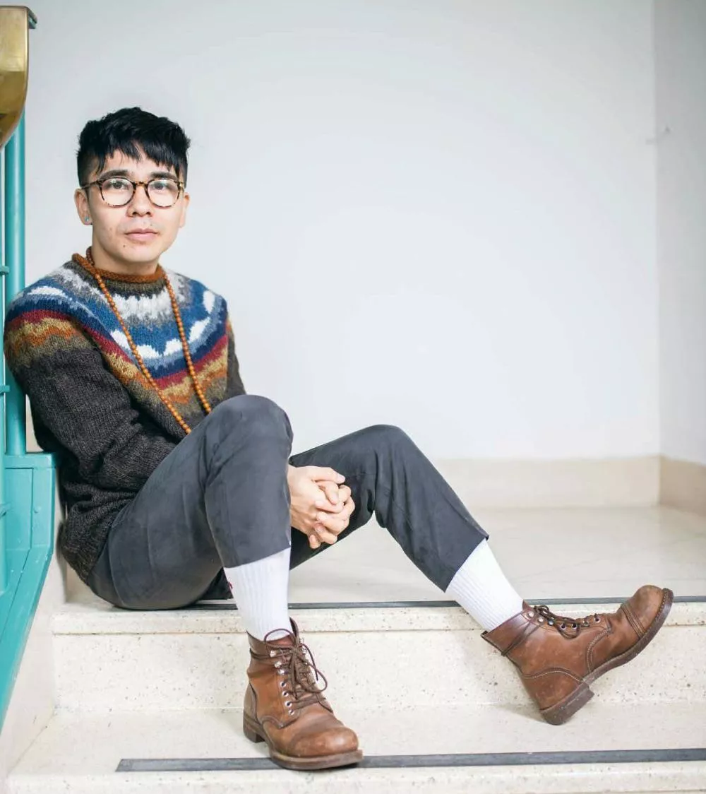 Un homme assis sur des marches, portant des lunettes, un pull coloré, un collier, un pantalon sombre, des chaussettes blanches et des bottes marron.