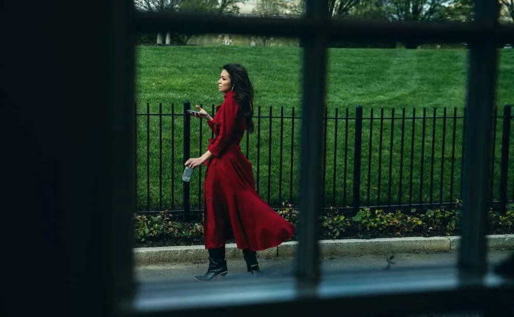 Une femme en robe rouge marche le long d'une clôture noire, tenant un téléphone et une bouteille d'eau, avec un espace vert en arrière-plan.