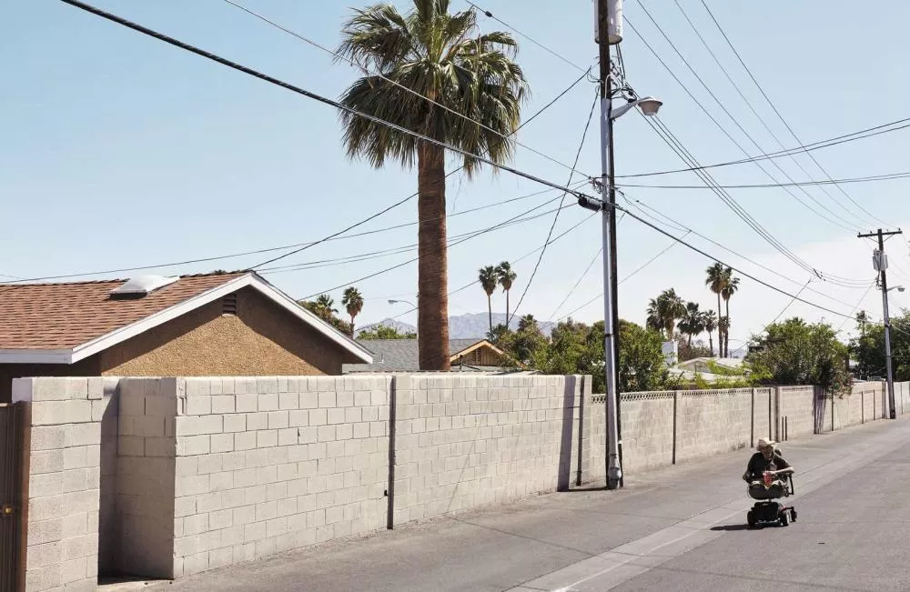 Une rue bordée de murs en parpaings, avec des poteaux électriques et des palmiers en arrière-plan. Une personne est assise sur un scooter électrique.