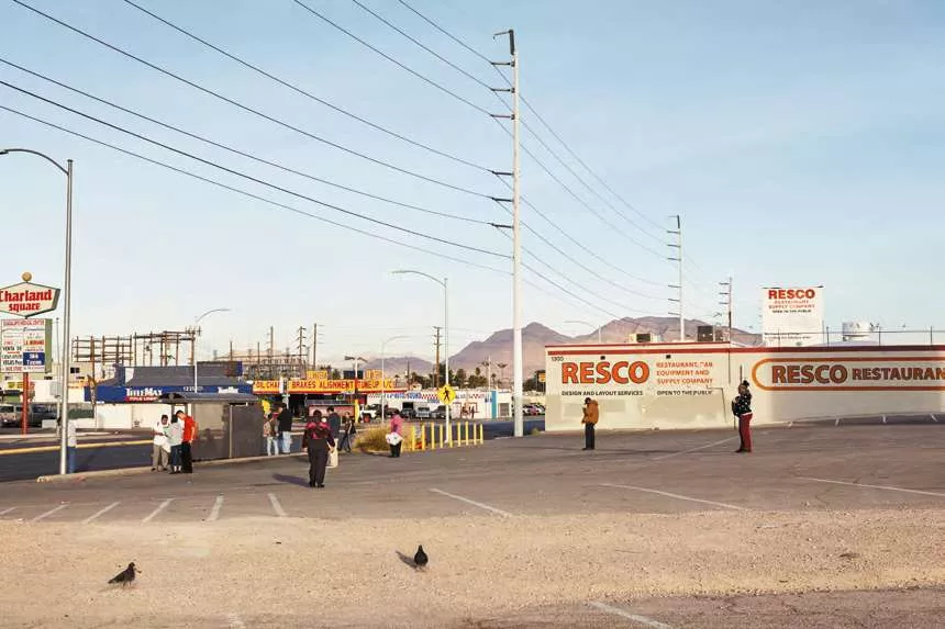 L'image montre un parking désert avec quelques personnes dispersées. À l'arrière-plan, on voit des bâtiments commerciaux, des panneaux publicitaires, et des montagnes à l'horizon.