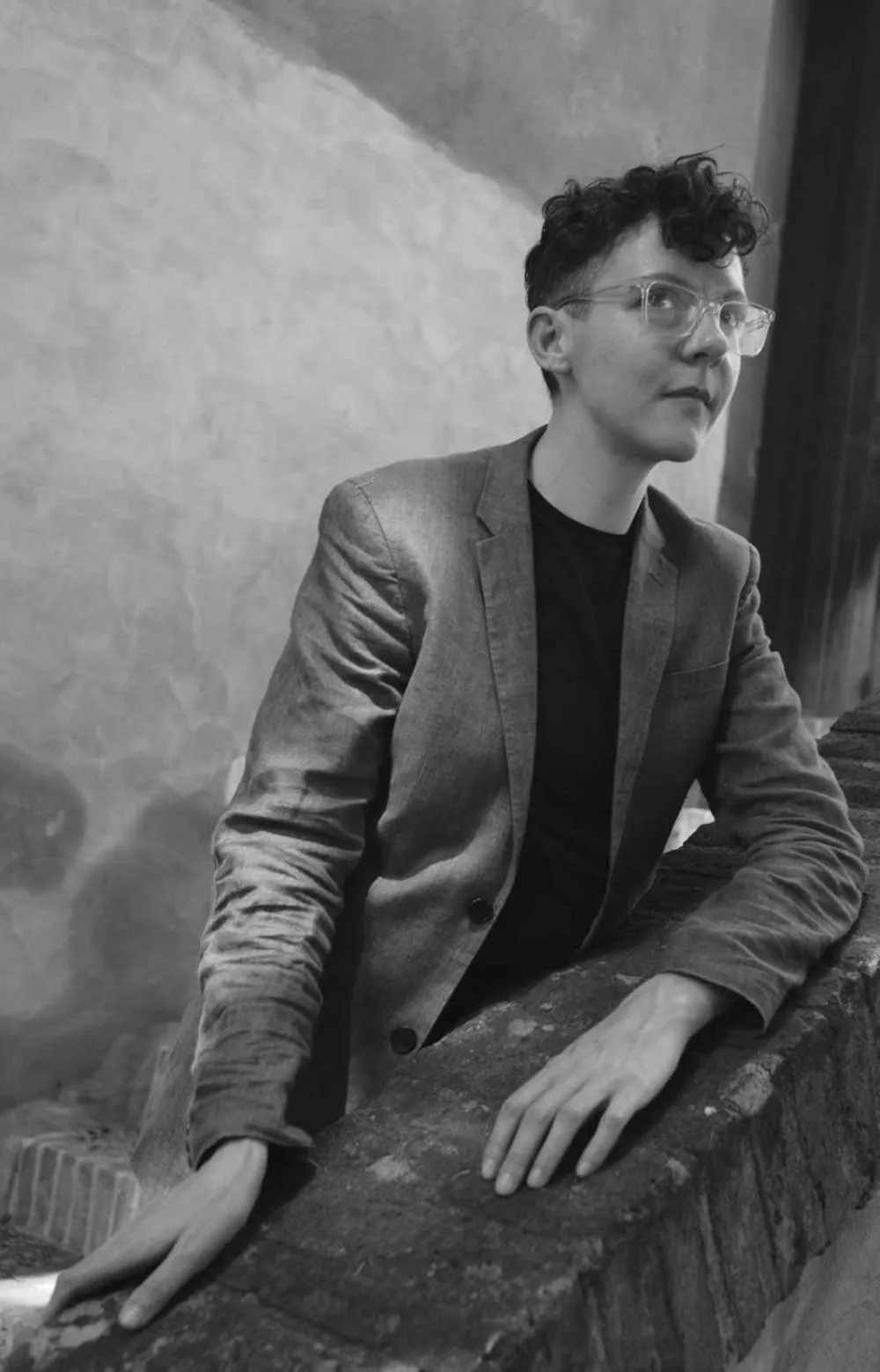 Une personne avec des lunettes et des cheveux courts bouclés, portant une veste, est appuyée sur un mur en briques dans un environnement extérieur. L'image est en noir et blanc.
