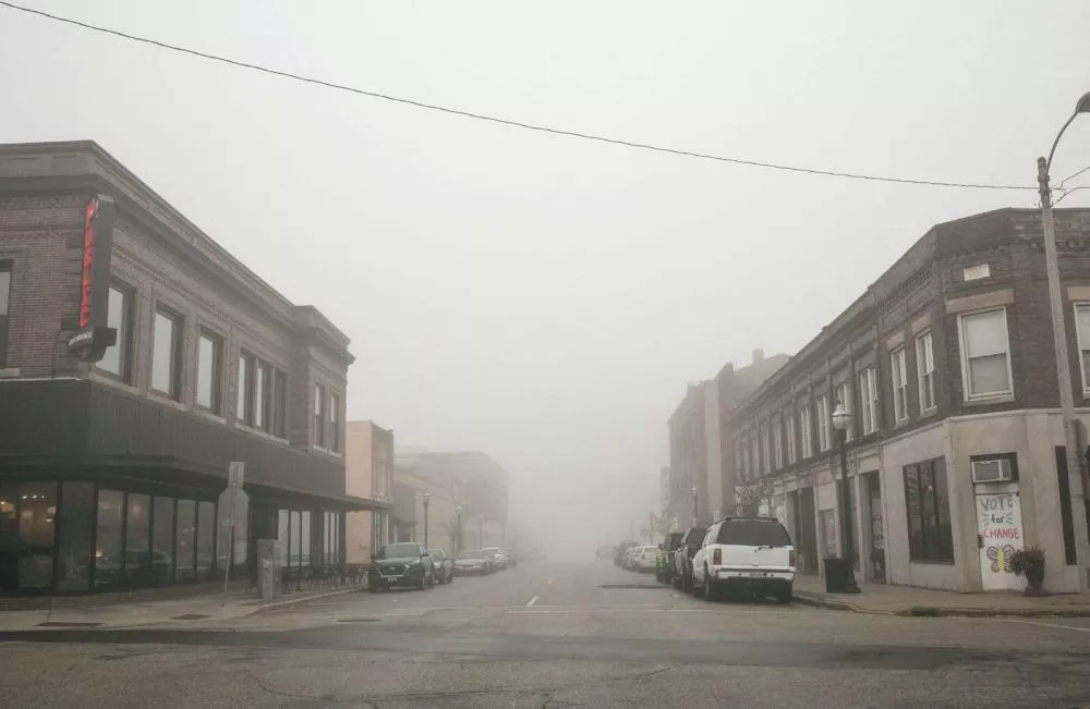 Une rue urbaine sous un épais brouillard, avec des bâtiments en briques de chaque côté et des voitures garées le long du trottoir. Un panneau sur un bâtiment indique "Vote for Change".