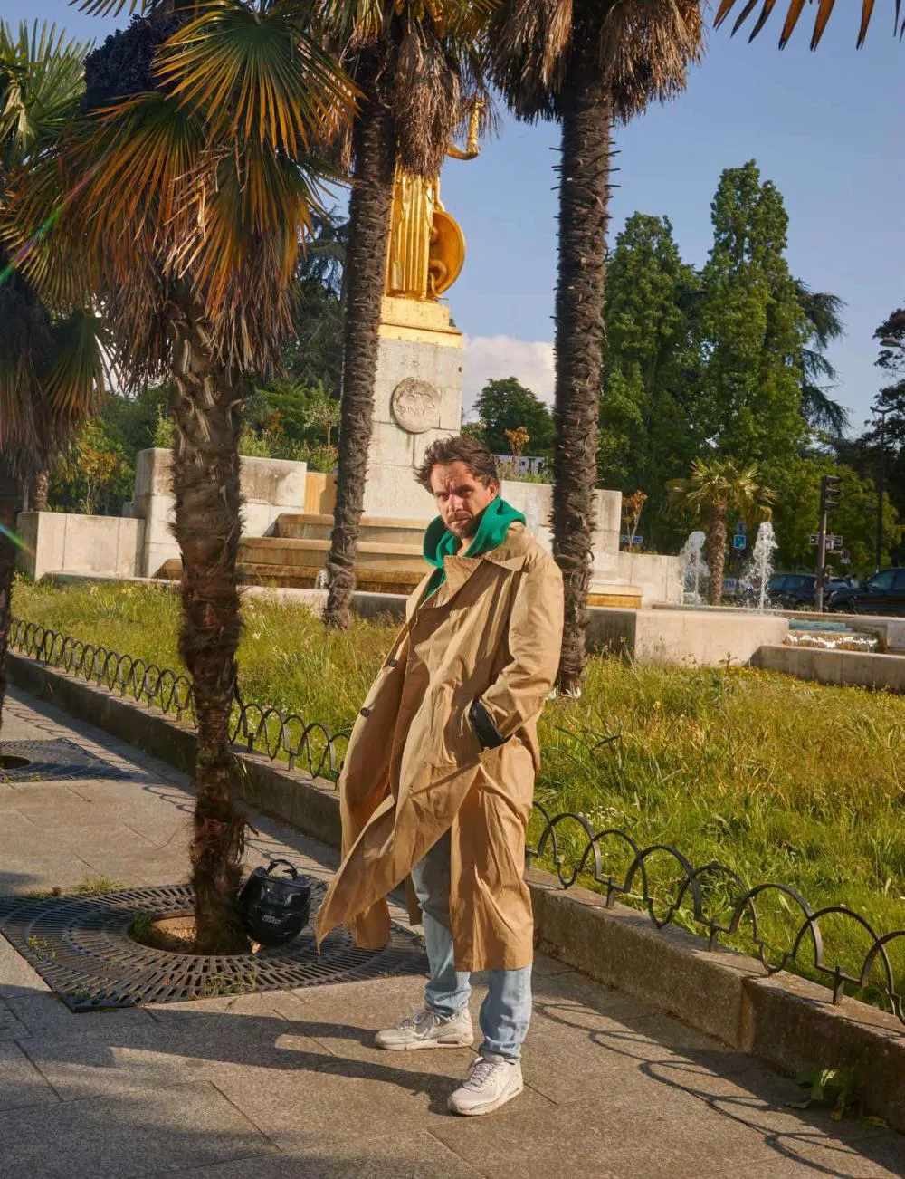 Un homme portant un long manteau beige et un sweat à capuche vert se tient debout sur un trottoir près de palmiers et d'une fontaine. Il a les mains dans les poches et il y a un sac noir posé près de lui.