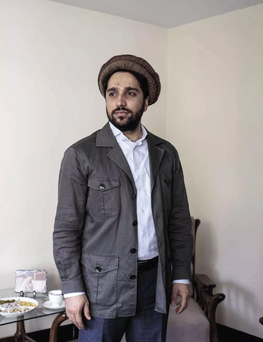 Un homme portant une veste grise et un chapeau traditionnel se tient debout dans une pièce. Il y a une table avec des tasses et une assiette à côté de lui.