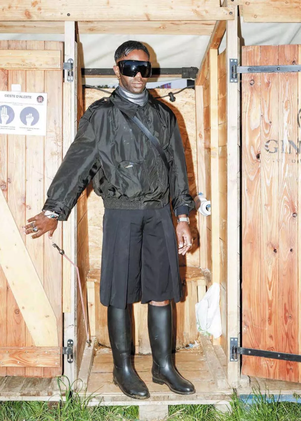 Un homme portant des lunettes de soleil, un blouson noir, un short ample et des bottes en caoutchouc se tient dans une cabane en bois.