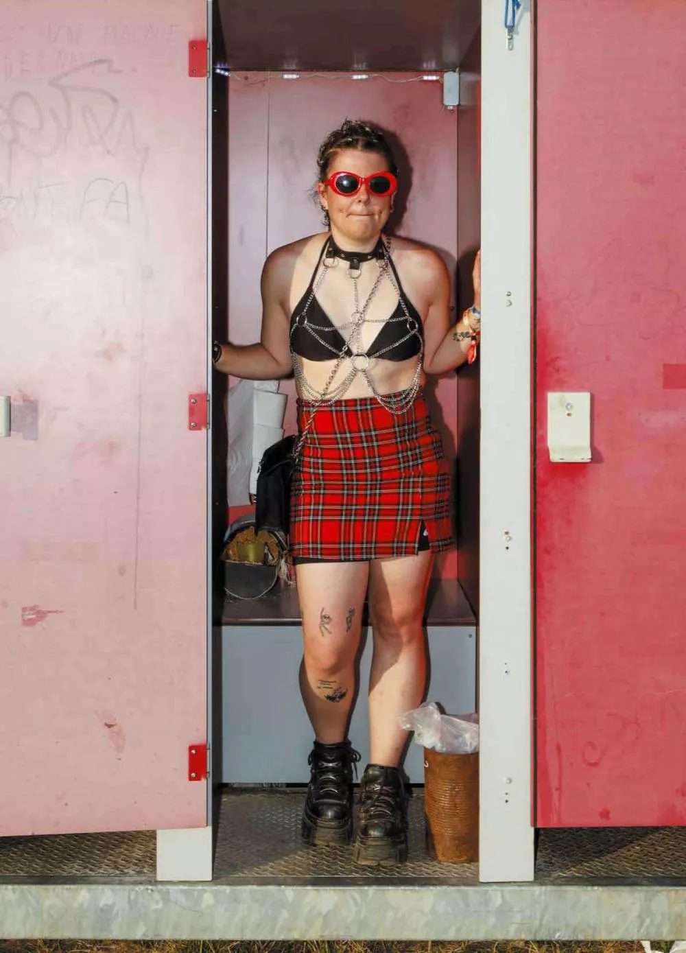 Une personne portant des lunettes rouges, un haut noir avec des chaînes, une jupe à carreaux rouges et noirs, et des bottes noires, se tient dans l'ouverture d'une cabine rose.