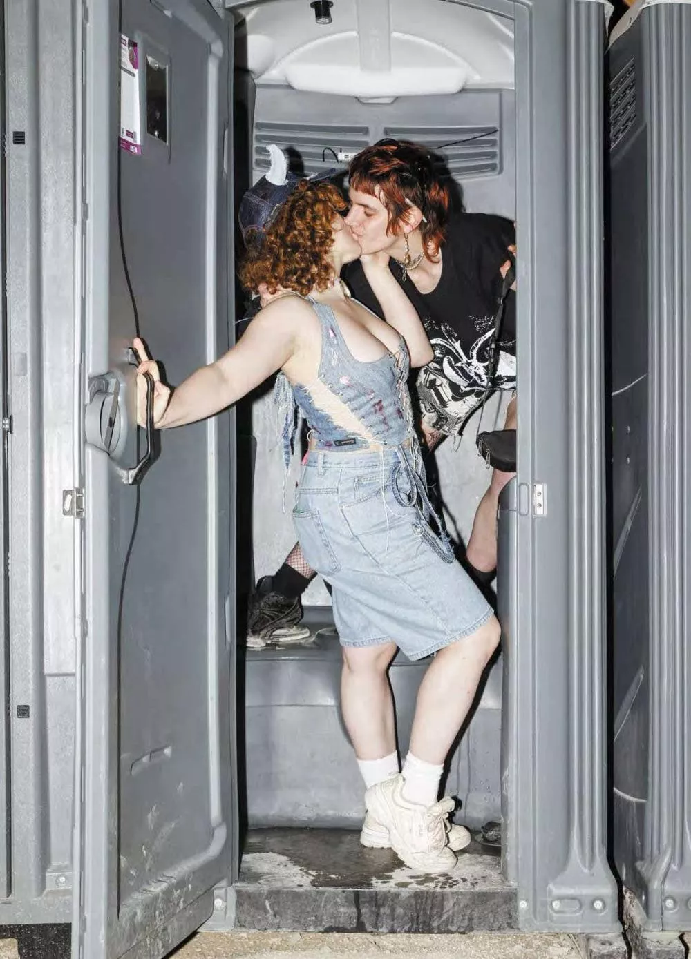 Deux personnes s'embrassent à l'intérieur d'une cabine de toilettes portatives. L'une porte une salopette en jean et des baskets blanches, tandis que l'autre est habillée en noir.