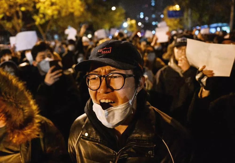 Un groupe de personnes manifestent la nuit, certaines portant des masques. Une personne au premier plan semble crier.