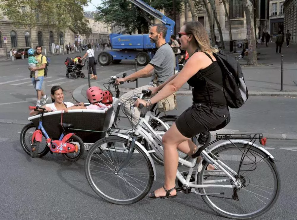Des personnes circulent à vélo dans une rue de la ville. Une femme conduit un vélo avec un bac à l'avant transportant un enfant portant un casque rouge. Un homme et une autre femme à vélo les accompagnent. D'autres personnes et un camion sont visibles en arrière-plan.