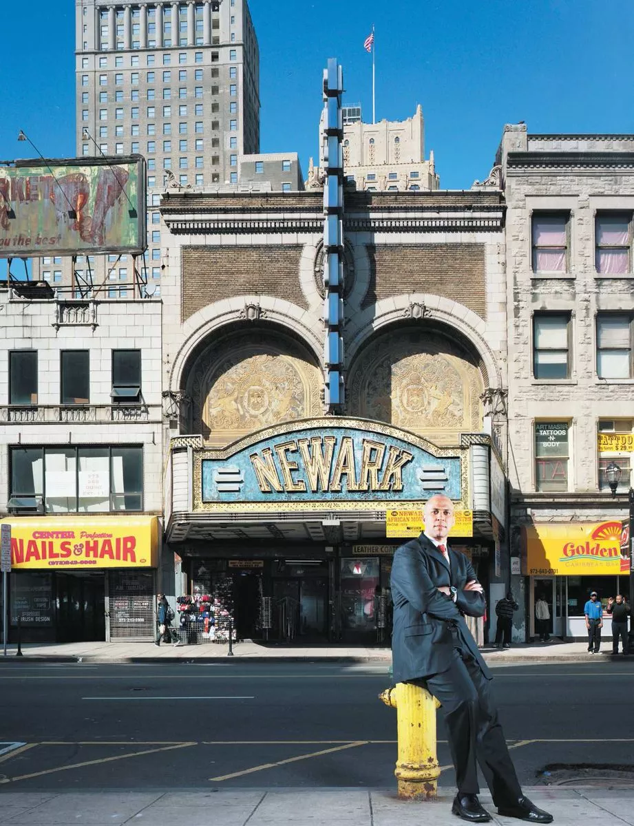Un homme en costume s'appuie sur une borne d'incendie devant un bâtiment arborant une enseigne "NEWARK". L'architecture est ornée et il y a des commerces de chaque côté, avec des enseignes visibles pour un salon de coiffure et un restaurant. En arrière-plan, on voit des immeubles et un drapeau américain.
