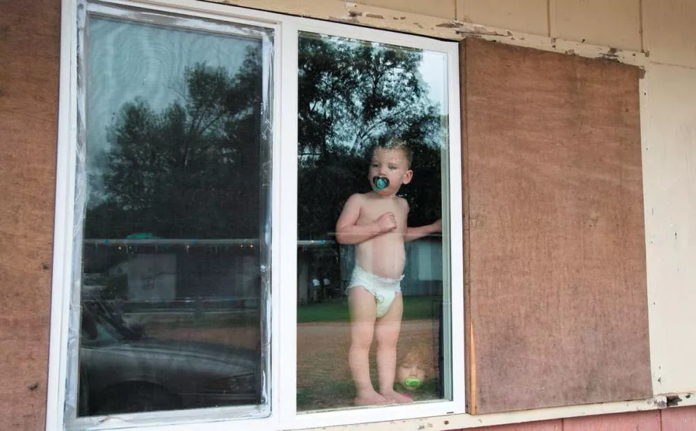Un enfant en couche avec une tétine se tient derrière une fenêtre, regardant à l'extérieur.
