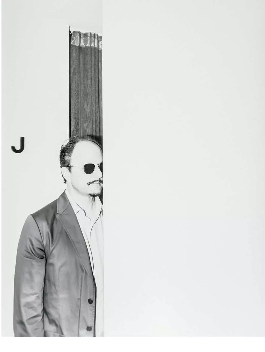 Un homme portant des lunettes de soleil et un costume se tient partiellement caché derrière un rideau, avec la lettre "J" visible à côté de lui.