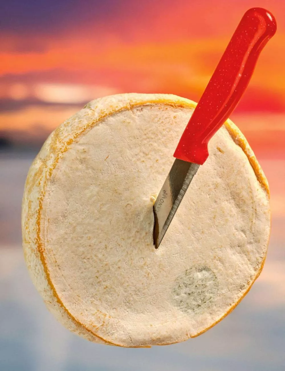 Une meule de fromage avec un couteau à manche rouge planté dedans, sur un fond coloré de coucher de soleil.