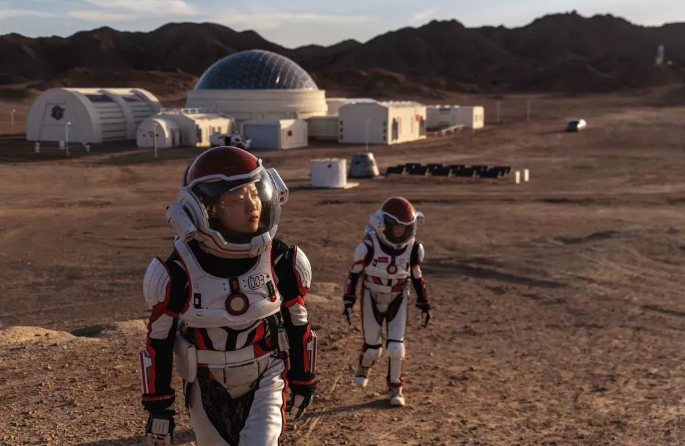 Deux personnes en combinaison spatiale marchent sur un terrain désertique, avec des installations futuristes en arrière-plan et des montagnes à l'horizon.