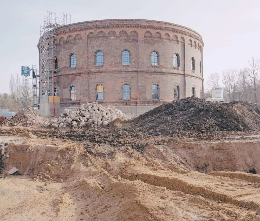 Un bâtiment en briques de forme cylindrique est en cours de rénovation, entouré d'échafaudages. Devant, il y a des tas de terre et de gravats, indiquant un chantier de construction.