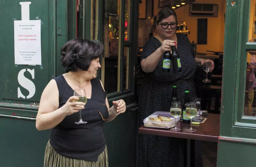 Deux femmes sont à l'entrée d'un établissement. L'une tient deux bouteilles de bière et un verre, tandis que l'autre tient un verre de vin blanc. Sur une table à côté d'elles, il y a des verres de vin blanc et un plateau de nourriture. Une affiche sur le mur mentionne une vente à emporter de grignotages maison.