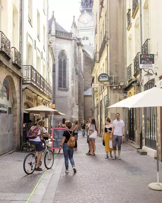 Une rue étroite et pavée avec des bâtiments anciens de chaque côté. Des personnes marchent ou font du vélo. Il y a des enseignes de magasins et des terrasses avec des parasols. Au fond, on aperçoit une église avec une grande horloge.