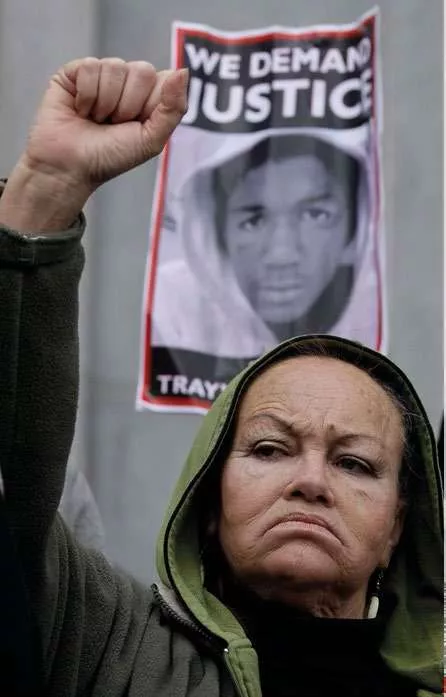 Une femme lève le poing en signe de protestation devant une affiche demandant justice, avec la photo d'un jeune homme en arrière-plan.
