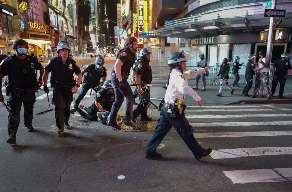 Des policiers en uniforme, certains portant des casques, sont en train de procéder à une arrestation sur un passage piéton dans une zone urbaine éclairée par des néons. Une personne est au sol entourée par plusieurs agents. D'autres personnes se trouvent à l'arrière-plan.