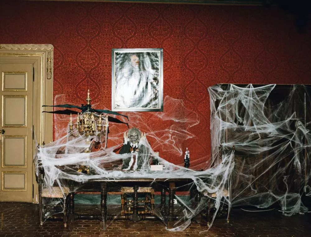 Une pièce décorée pour Halloween avec des toiles d'araignée couvrant un chandelier, une table et un meuble. Un squelette décoratif est posé sur la table. Les murs sont rouges avec un motif, et un tableau est accroché au mur.
