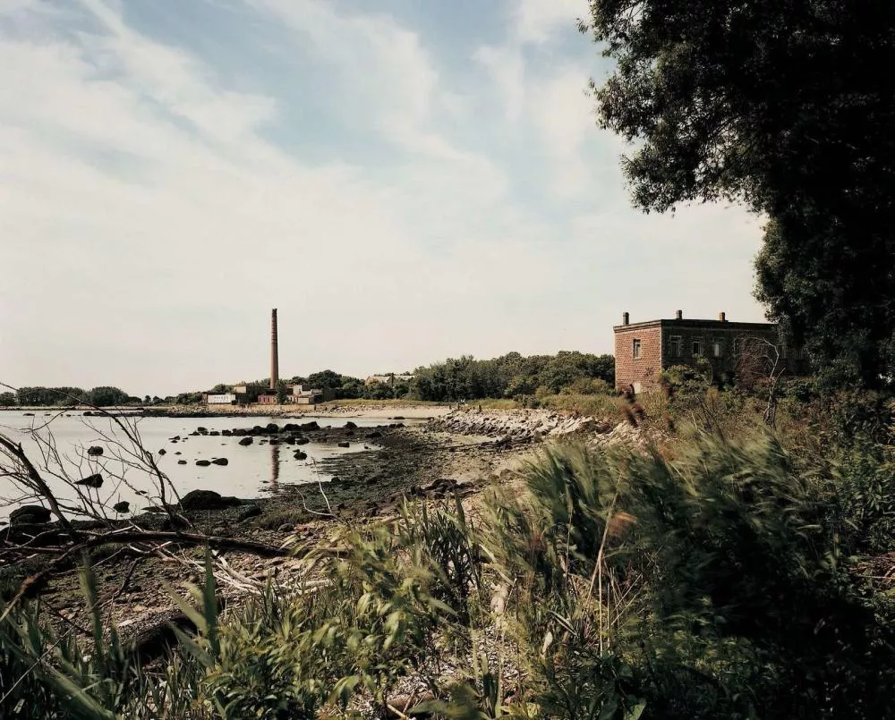 Une scène côtière avec un bâtiment en briques à droite, des arbres environnants et une cheminée industrielle au loin. Le rivage est parsemé de rochers et de végétation.