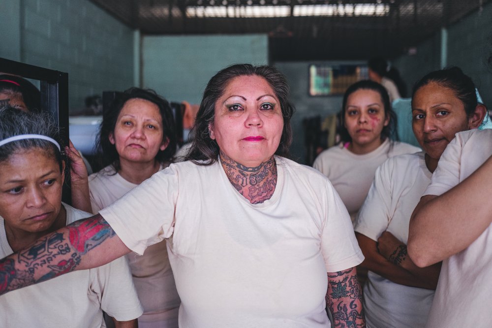 Un groupe de femmes en t-shirts blancs, certaines avec des tatouages, se tient dans une pièce. La femme au centre a des tatouages visibles sur le cou et le bras. Elles semblent être dans un environnement intérieur avec des murs en briques.