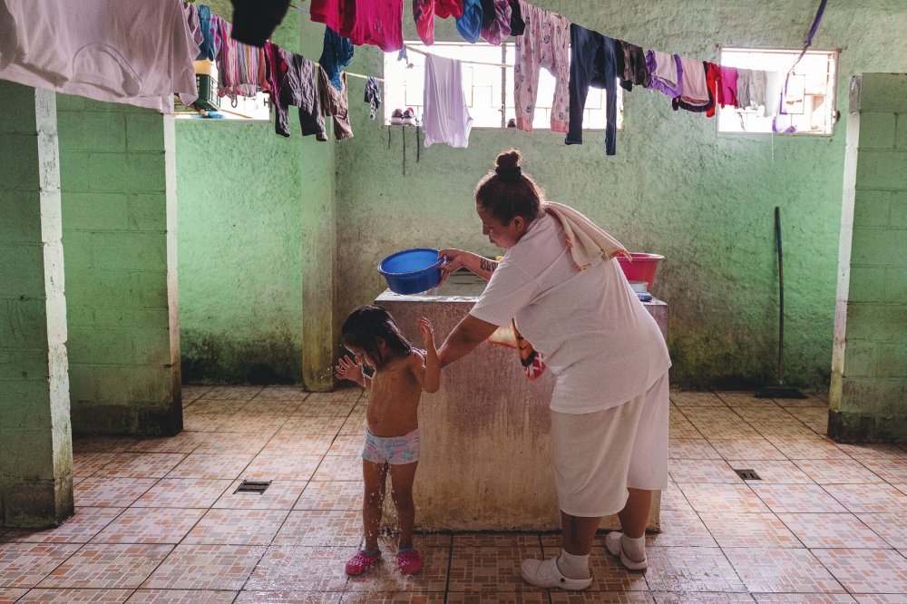 Une femme verse de l'eau sur une petite fille dans une pièce avec des vêtements suspendus pour sécher. La pièce a des murs verts et un sol carrelé.