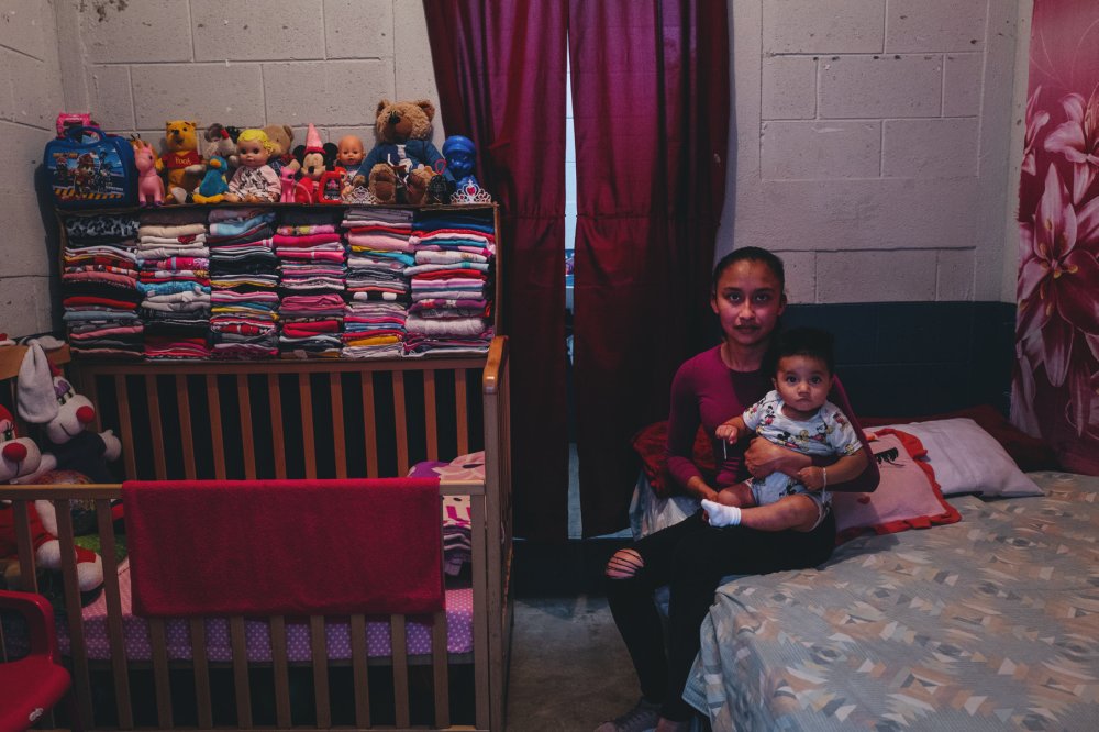 Une femme est assise sur un lit, tenant un bébé. À gauche, il y a un lit d'enfant avec des jouets en peluche et une étagère remplie de vêtements pliés et de poupées. Les rideaux sont rouges et le mur est en béton.