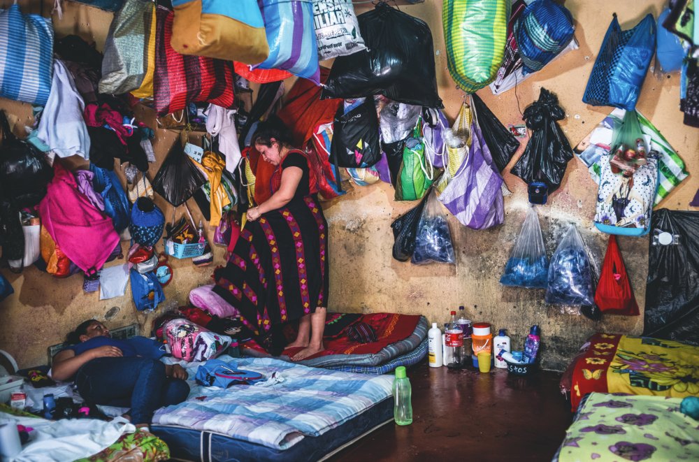 Une pièce encombrée où des sacs colorés sont accrochés aux murs. Deux personnes se trouvent à l'intérieur, l'une allongée sur un matelas et l'autre debout. Des objets personnels et des bouteilles sont dispersés autour.