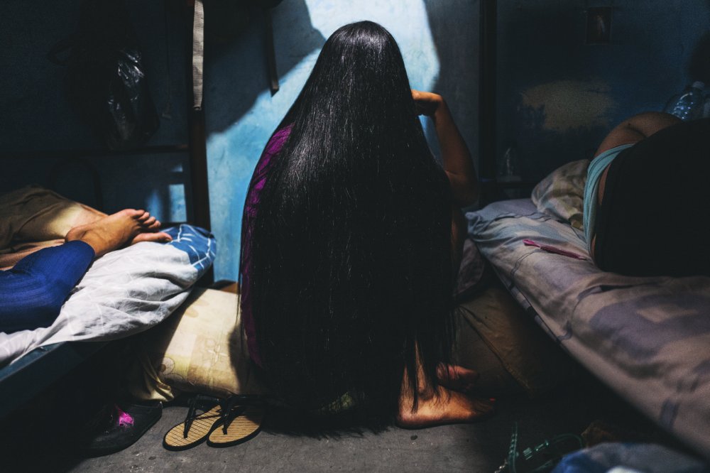 Une personne aux longs cheveux noirs est assise sur un lit dans une pièce sombre. On voit d'autres personnes allongées sur des lits autour d'elle, et des sandales posées au sol.