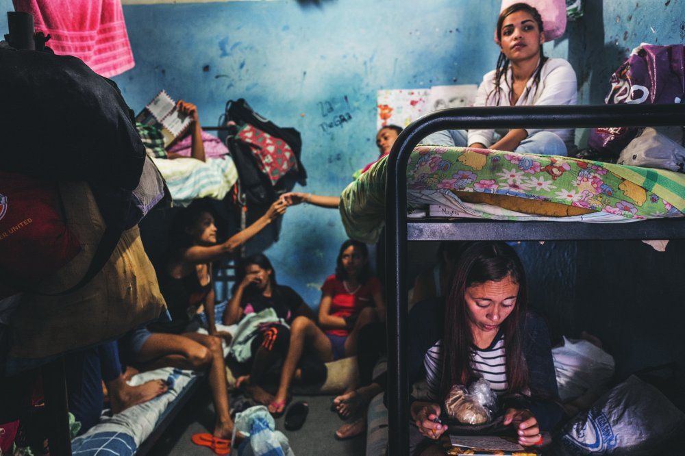 Un groupe de femmes est rassemblé dans une petite pièce avec des lits superposés. Certaines sont assises ou allongées sur les lits, tandis qu'une femme est assise par terre, lisant ou regardant quelque chose. L'environnement est encombré avec des sacs et des vêtements.