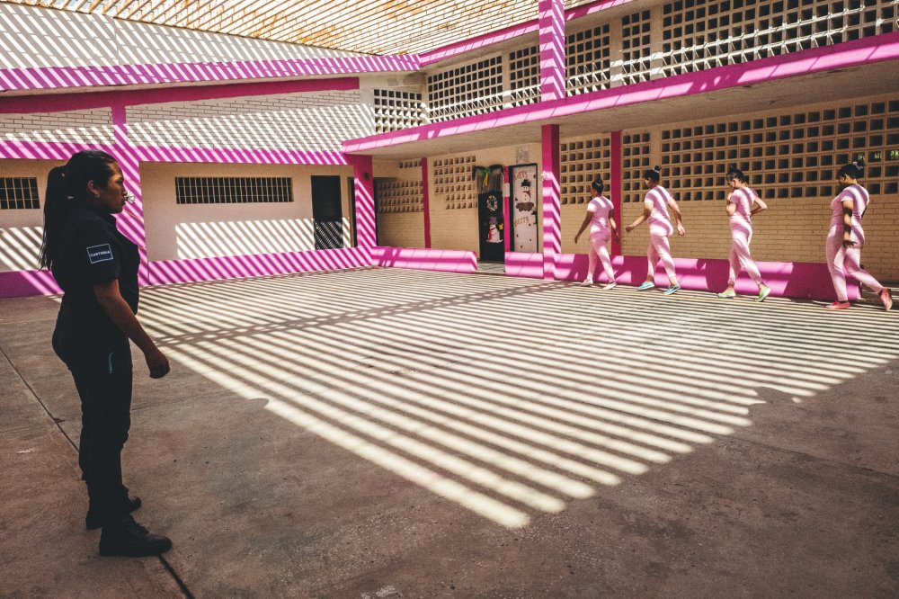 Une femme en uniforme surveille quatre femmes en tenue rose qui marchent en file indienne dans une cour, avec des ombres de lignes horizontales projetées sur le sol et les murs, qui sont partiellement peints en rose.