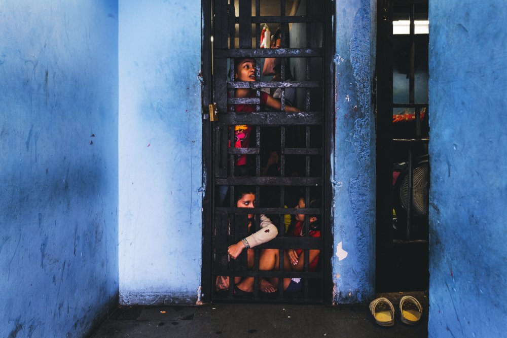 Des enfants sont assis derrière une porte en métal à barreaux, dans une pièce aux murs bleus. Deux paires de chaussures sont posées à l'extérieur de la porte.