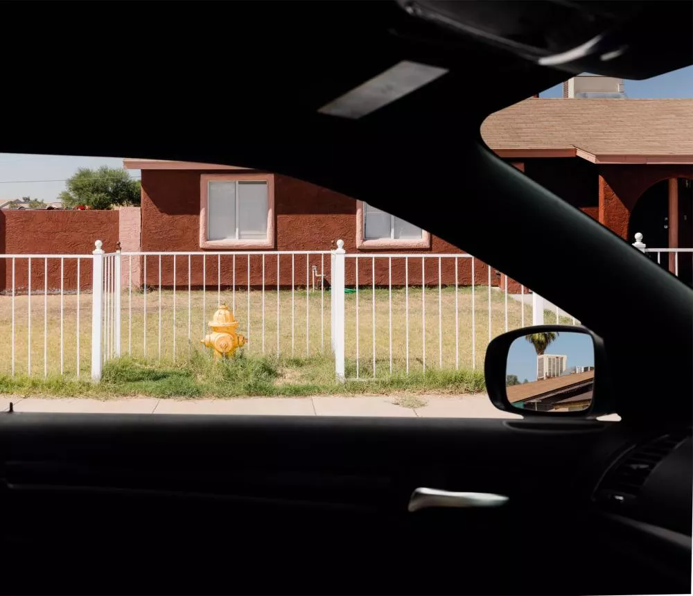 Vue depuis l'intérieur d'une voiture, on aperçoit une maison en briques rouges avec une clôture blanche et une bouche d'incendie jaune sur la pelouse. Dans le rétroviseur, on voit un bâtiment en arrière-plan.