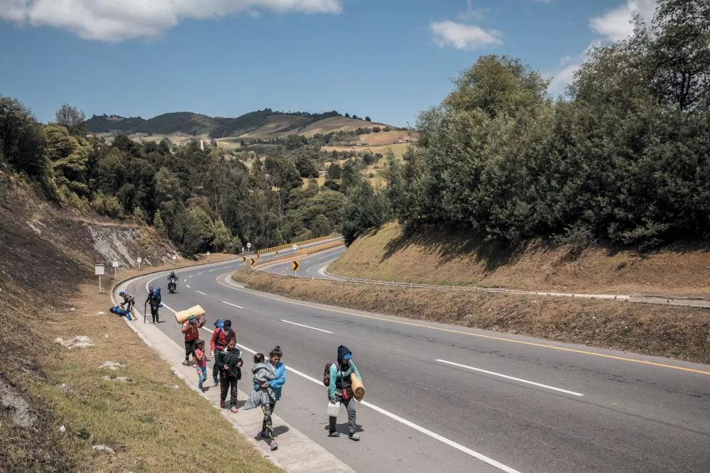Un groupe de personnes marche le long d'une route sinueuse dans un paysage vallonné et verdoyant. Certaines portent des sacs à dos et des affaires, tandis que deux motos circulent sur la route.