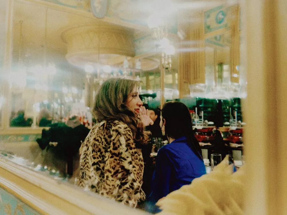 Deux personnes sont assises dans un café ou un restaurant élégant. L'une porte un manteau à motif léopard et l'autre un vêtement bleu. L'ambiance est chaleureuse avec des lumières douces et des miroirs ornés.