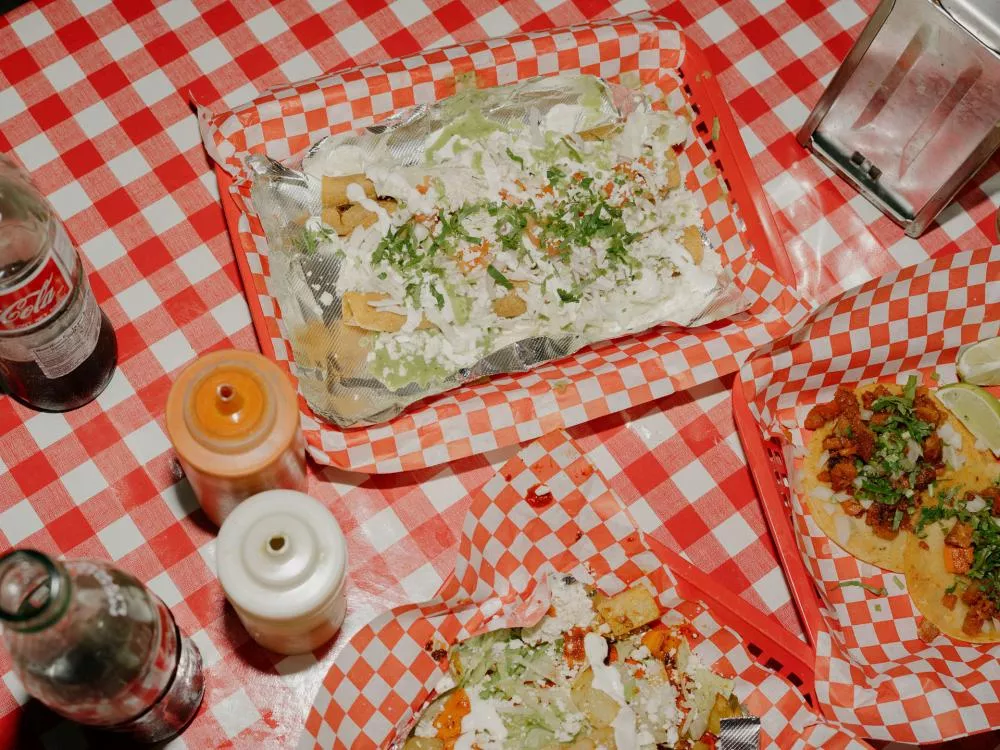 Un repas composé de tacos garnis de viande, de fromage, de crème et de coriandre, accompagné de sauces dans des bouteilles et d'une bouteille de soda, le tout sur une nappe à carreaux rouges et blancs.
