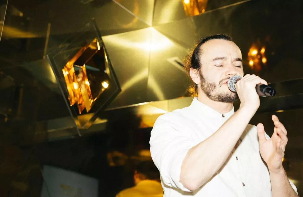 Un homme chante dans un microphone, les yeux fermés, dans un environnement lumineux avec des reflets dorés.