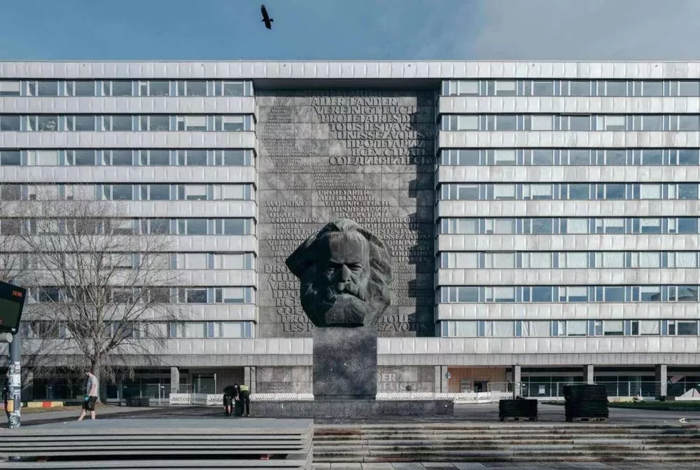 Un bâtiment moderne avec une grande sculpture en relief d'un visage barbu au centre, entourée de texte en plusieurs langues. Une personne marche sur le trottoir et un oiseau vole dans le ciel.